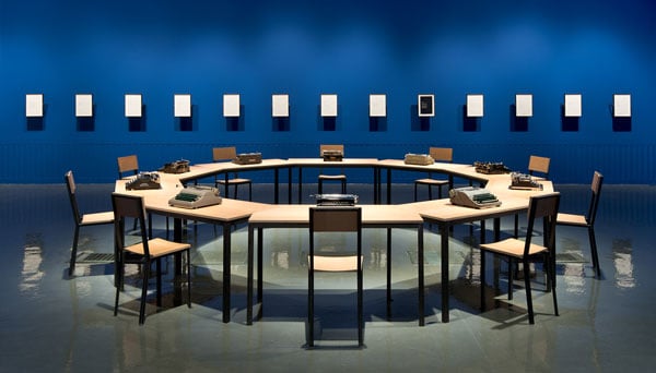 A photograph of an art installation shows a circular table surrounded by 10 chairs. At each place sits a manual typewriter. A facing wall is painted a sapphire blue.