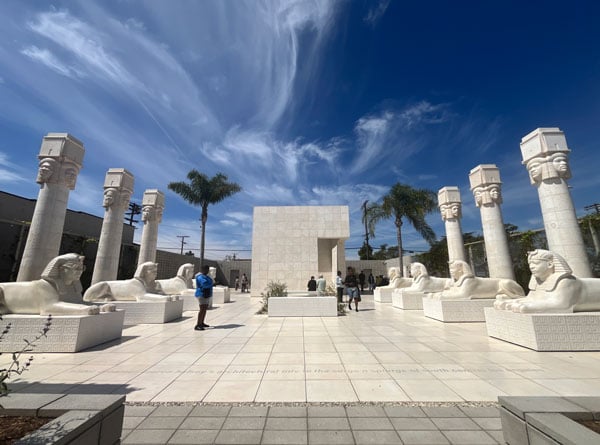 A view of a sculpture park by Lauren Halsey features two rows of sphinxes and two rows of Hathoric columns flanking a gleaming white courtyard under a swirl of clouds.