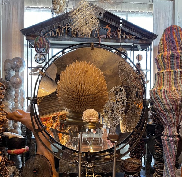 A photograph of a cluttered gallery spaces shows a large convex mirror surrounded by curious objects, including a Classical pediment that hangs from above.