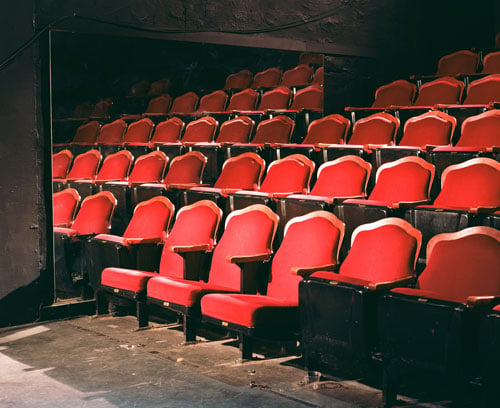 A color photograph shows the interior of a small theater lined with crimson red theater seats trimmed in wood.
