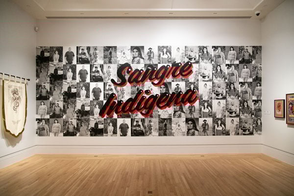 A wall mural in a gallery features a collage of black and white portraits of people topped by the painted phrase, "Sangre Indigena."