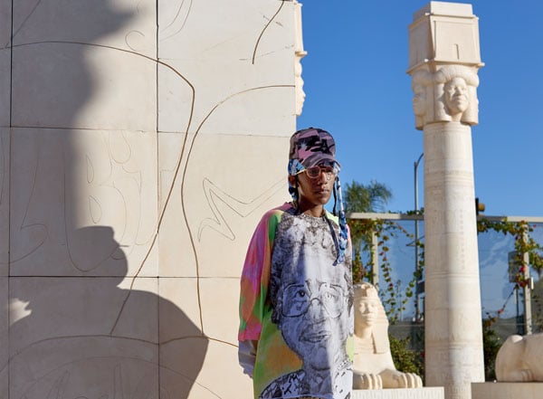 The artist Lauren Halsey, dressed in a brightly colored shirt and camo baseball cap, stands in front of a bas relief at her "sister dreamer" park in South LA.