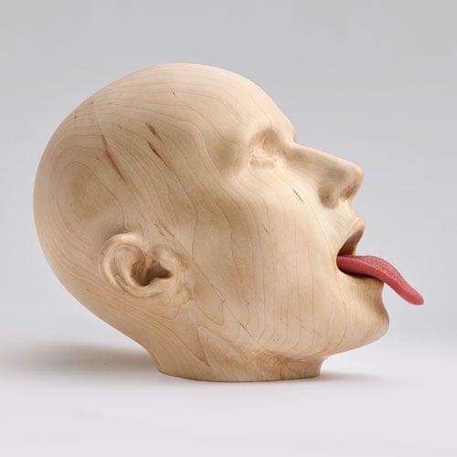 A wooden sculpture of a human head shows an androgynous figure sticking out their tongue.