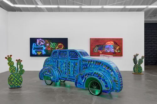 An installation view of a gallery shows a whimsical wooden sculpture of a car illuminated by green neon — and framed by sculptures of cactus.