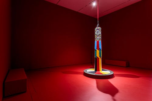 A beaded punching bag sculpture is dramatically illuminated inside a gallery painted a deep red.