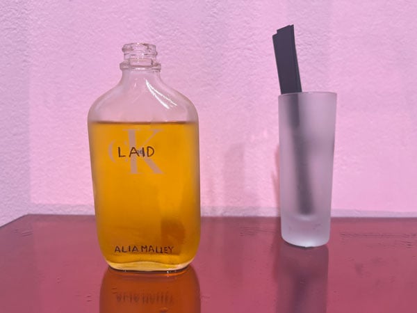 A bottle of perfume bearing the word "Laid" on the front sits on a gallery shelf next to a batch of scent strips