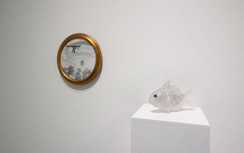 A circular graphite drawing shows a burned power line while in the foreground, a delicate cast glass sculpture of a translucent fish rests on a plinth.
