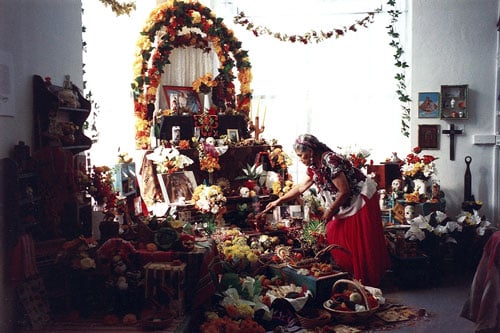 A color snapshot shows Ofelia Esparza, a Mexican woman in traditional dress, tending to a large Day of the Dead altar featuring flowers, photographs and fruit.