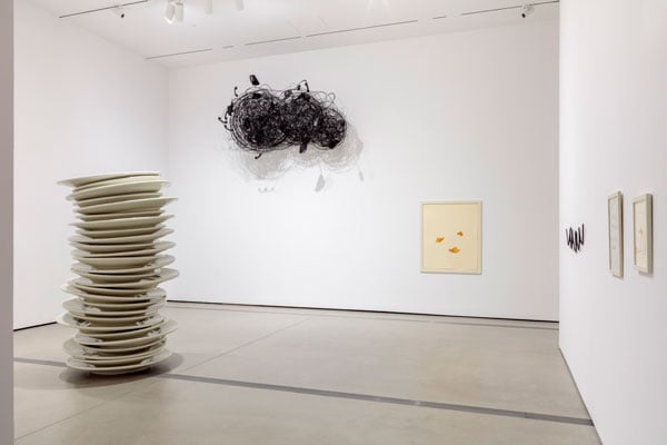An installation view of Robert Therrien's retrospective shows a sculpture that resembles a towering stack of dinner plates, a wall piece made from a tangle of telephone wire, and various framed drawings.