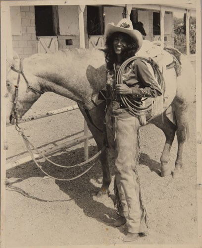 A black and white image shows a Black woman in chaps and a cowboy hat posing next to a white horse.