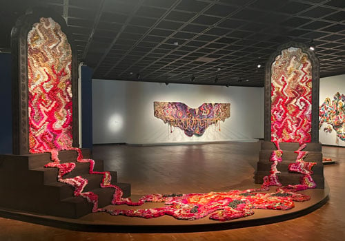 A textile installation by Suchitra Mattaiw shows a pink and red colored weaving that seems to spill and puddle from a pair of arched wooden doorways.