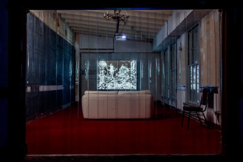 A black and white films screens in a small room with a white couch protected by a clear plastic curtain.