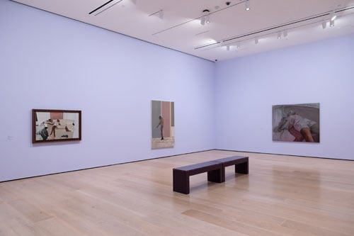 An installation view of Noah Davis's  survey at the Hammer museum show three paintings of figures installed against a lavender colored wall and a purple bench.