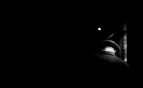 A black and white photo shows a man in a hat from behind at night, his head bowed down so his neck is visible.