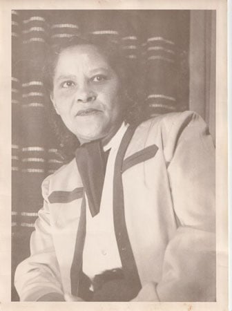 A vintage photographic portrait shows a fair-skinned Black woman wearing a stylish neckscarf and a suit trimmed with a band of dark fabric.