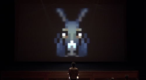A giant 8-bit head of a donkey looms on a screen over a person standing at a podium, their back turned to the viewer.