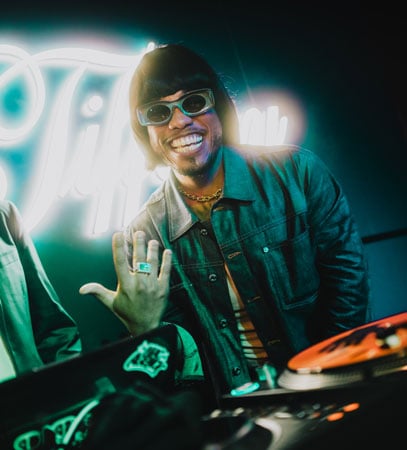 A portrait of DJ Pee .Wee (aka Anderson .Paak), shows the musician in sunglasses and a bob with bangs, smiling as he stands behind a turntable.