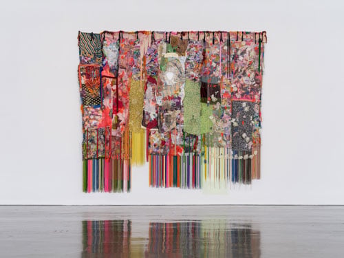 A brightly colored textile collage with multicolored fring at the bottom, by Elliott Hundley, hangs on a gallery wall at Regen Projects.