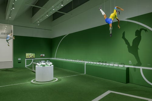 A view of a gallery shows the floor and walls decorated to resemble a soccer field. Dangling from the ceiling is a sculpture of a figure of a player in blue shorts and a yellow jersey.