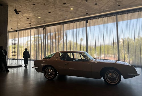 A sportscar in a museum gallery is silhouetted against daylight from floor to ceiling galleries.