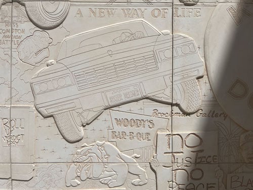 A bas relief by Lauren Halsey shows a lowrider, a sign for Woody's Bar-B-Que, a cartoon bulldog and a protestor holding up a sign that reads "No Justice No Piece"
