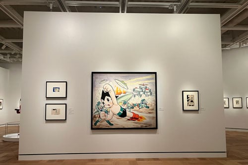 A view of a gallery wall shows a large painting of the Japanese character Astroboy hanging on a wall with three smaller pieces.