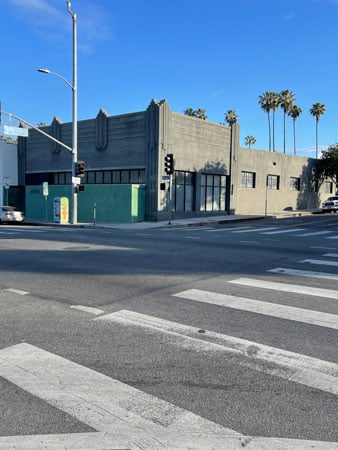 A dull green-grey building sits on an LA street corner. Behind it rise palm trees.