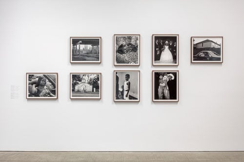 On a gallery wall hang eight black and white photographs by Rahim Fortune, including and image of a little boy, a farmhouse in a rural area, and a girl dressed up for a party.
