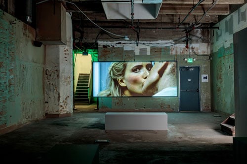 A video of two people kissing plays on a screen in a grimy basement-like space.