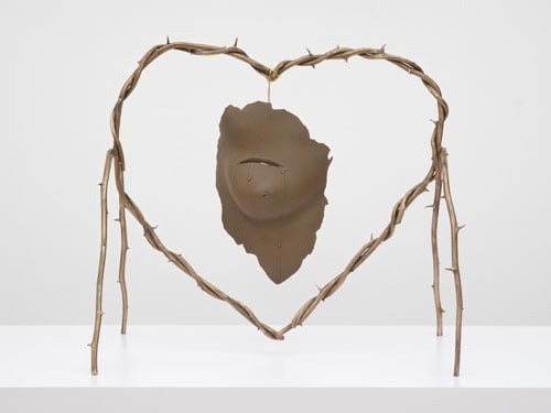 A bronze sculpture features a fragment of a human face — the chin and mouth — framed by a barbed wire heart.