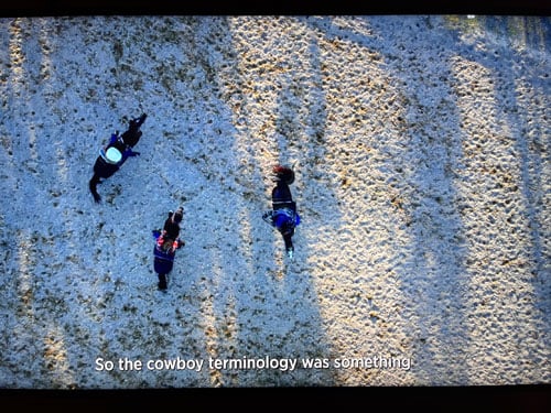 A still from a documentary shows a bird's eye view of people on horses on sandy terrain, with the subtitles "So the cowboy terminology was something..." at bottom