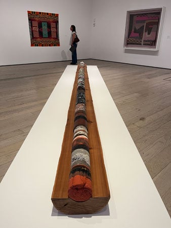 Half of a trunk of a cedar tree rests on a low-rise plinth in a gallery. It's core has been stuffed with plastic and other waste.
