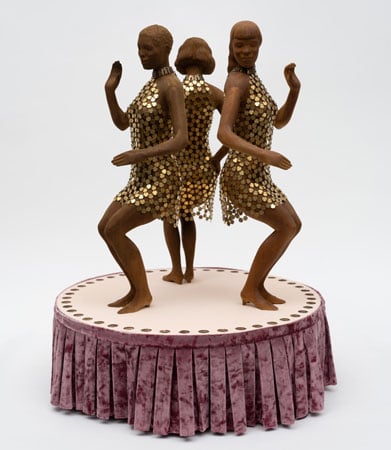 A sculpture made from brass, velvet, nylon and acrylic shows three Black women in sparkling gold dresses dancing on a small round stage.