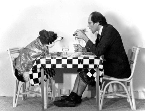 A film still shows a man a dark suit jacket, culottes and eyeliner sitting at a dinner table with a dog wearing a sparkling cape.