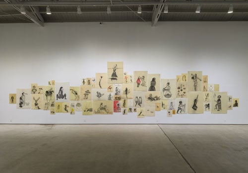 A salon style hang in a gallery features dozens of drawings — largely in black and white, though some bear splashes of red/