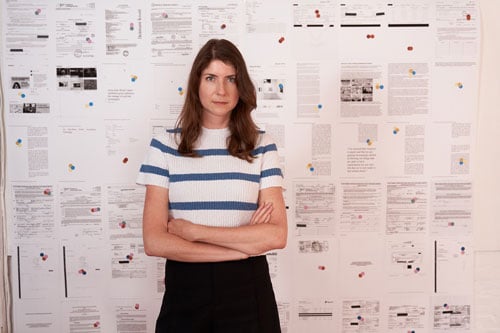 A female artist with long brown hair stands with her arms crossed before a wall papered over with documents.