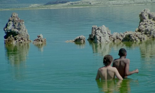 A still of a video by Stanya Kahn shows two boys — one white, one black — wading into a lake with rock formations.