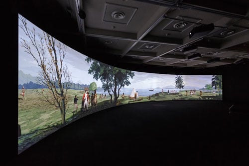 A darkened gallery shows a large curving screen that shows a large projection of people in a South Pacific landscape