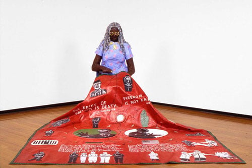 A paper mache sculpture by Willie Birch shows a Black woman with long grey hair stitching a quilt that describes aspects of African American history.
