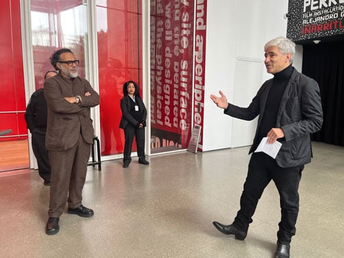 Filmmaker Alejandro Iñárritu stands with his arms crossed in a brown suit as he watching LACMA director Michael Govan talking.