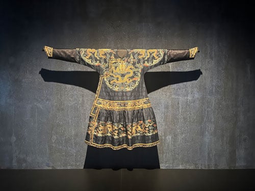 An 18th man's robe from China with gilded details is presented with arms extended to the sides in a gallery space stained a deep grey.