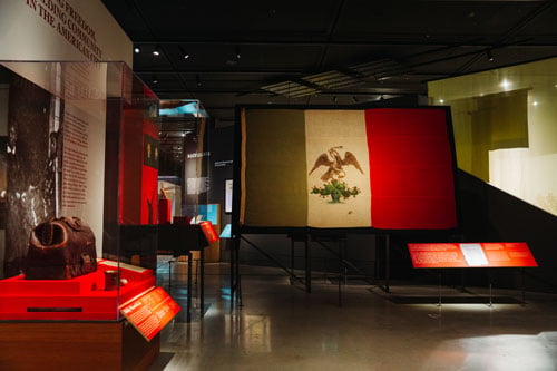 A handpainted Mexican flag is displayed in a dramatically lit gallery