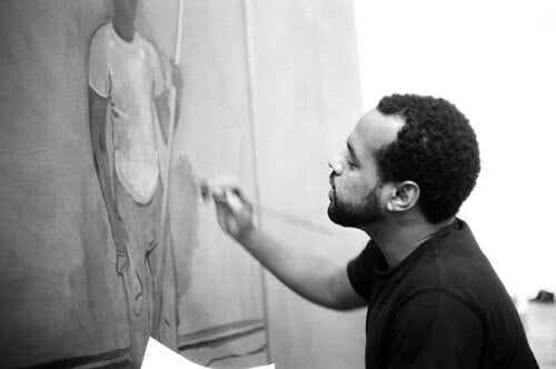 A black and white image shows Noah Davis, a Black man, painting a figure on a large canvas.