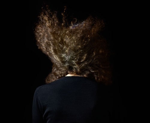 A moody color photograph by Eileen Cowin shows a woman with curly hair, photographed from behind, her hair looking as if it is being whipped by a gust of wind.