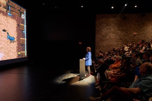 A person in a bright blue coat stands at a podium before an audience manipulating controls of a video game featuring donkeys on a screen.