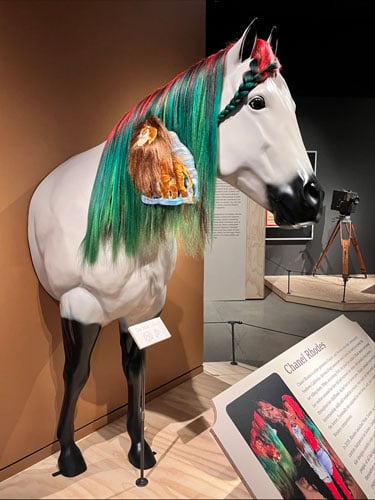 A gallery displays a sculpture of a horse wearing a green and pink mane.
