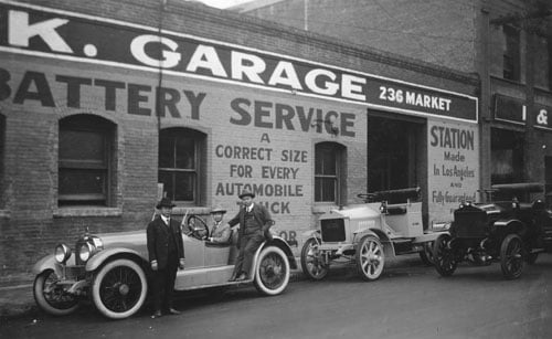 A vintage black and white photo from the 1910, shows three men in suits and hats posing before a car and a warehouse building that reads, "F&K Garage"