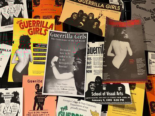 Posters by the Guerrilla Girls in bright yellow, black, white and red are tacked in an informal fashion to a gallery wall.
