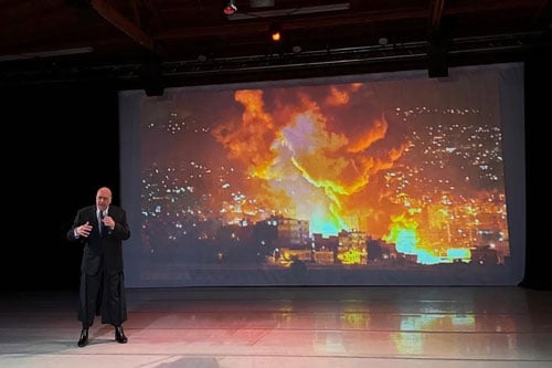 A man in a suit sings on stage before a video screen showing explosions in wartime.