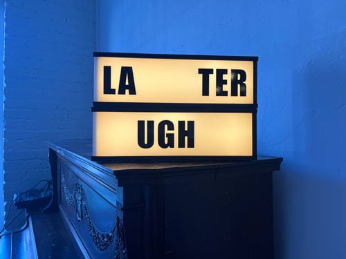 A sculpture in the form of a sign rests on top of a piano and can be read as "Later UGH" or "Laughter"